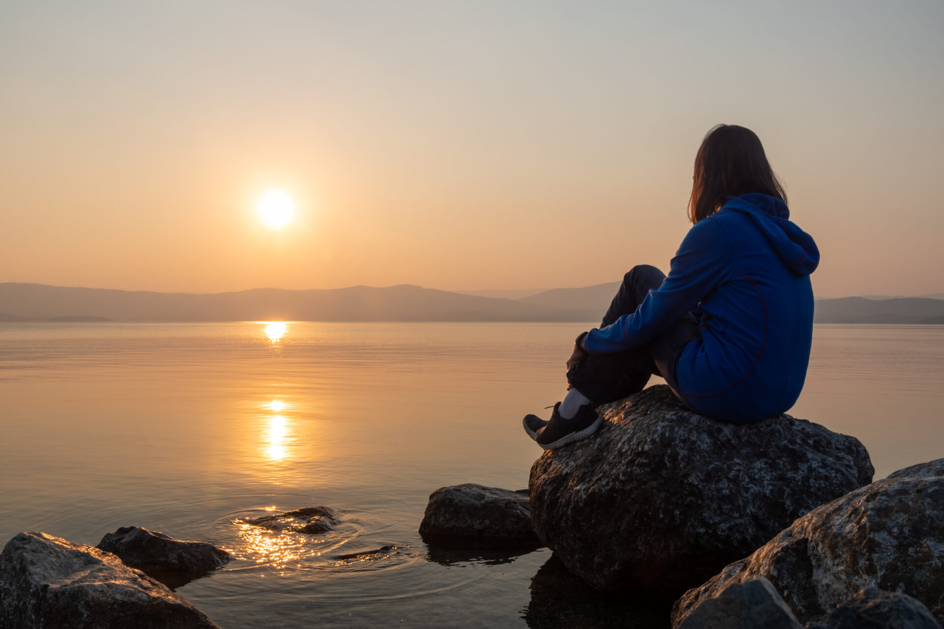 Girl looking sunset on sea, lake. Woman sitting on rocks. Lonely person. Girl alone outside. Vacation on sea